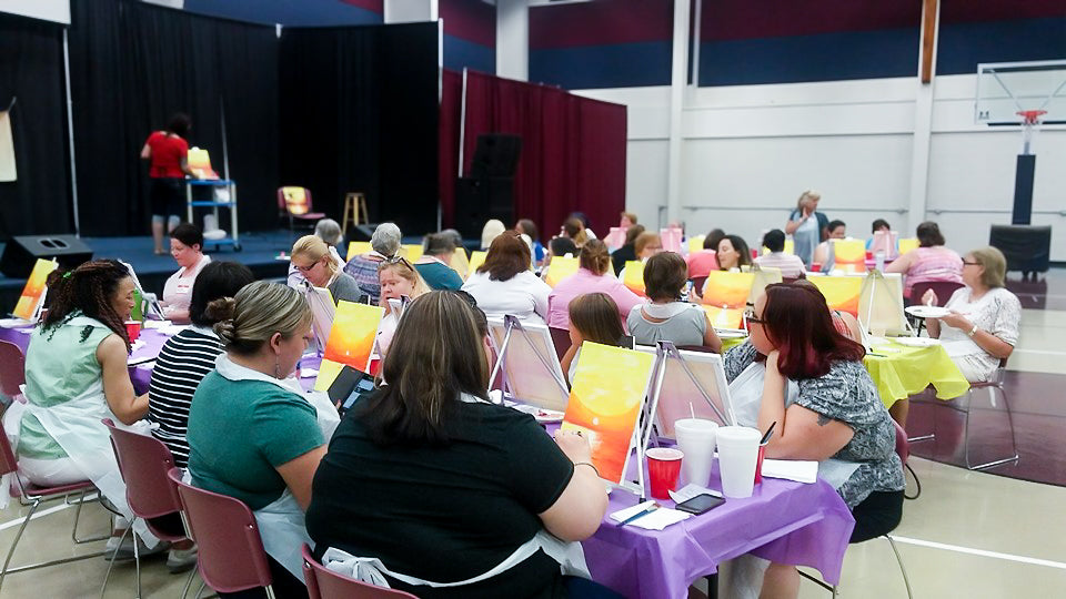 Large group of women at a painting even painting a palm tree sunset with the ocean in the background. Ladies are in a large church gym