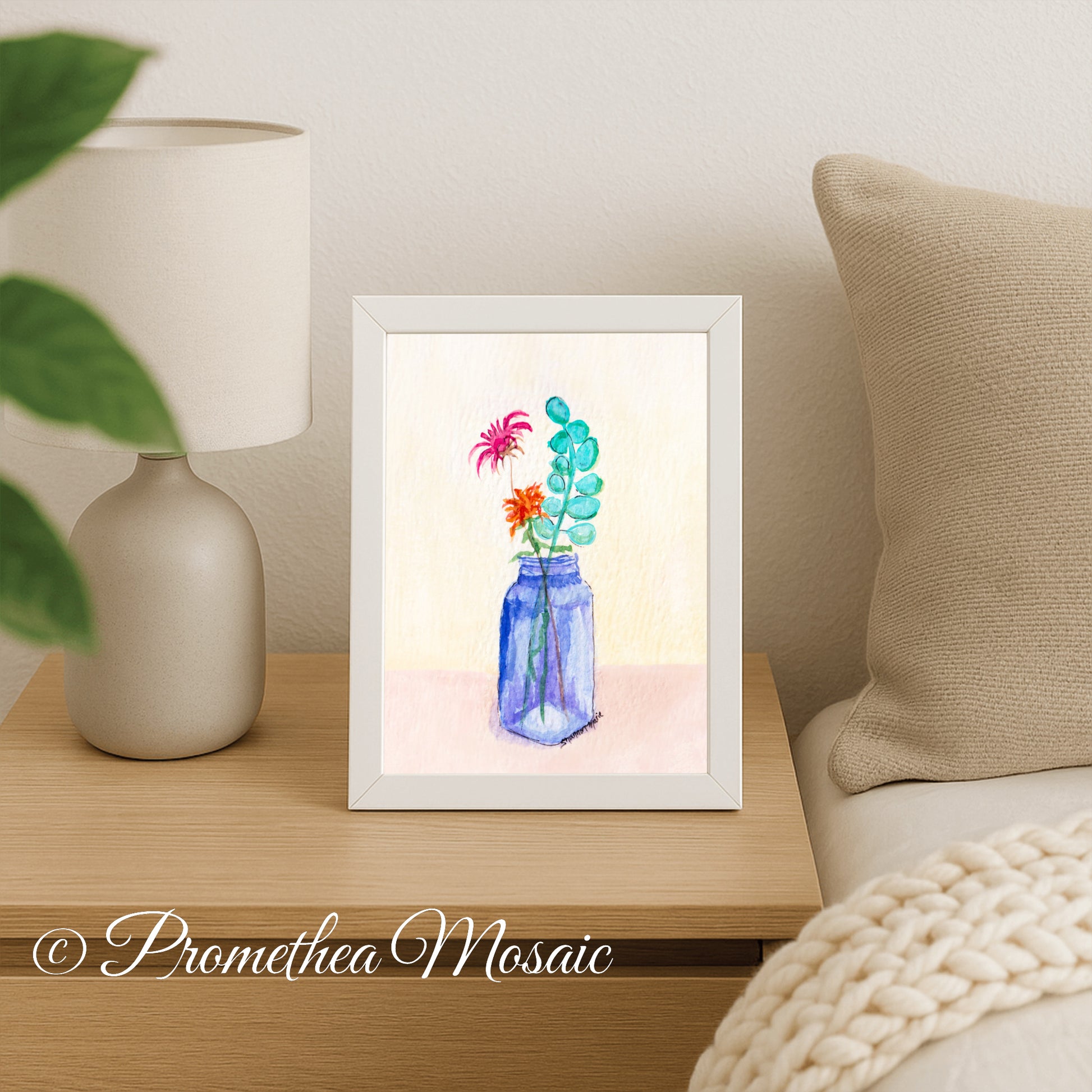 Framed artwork of flowers in a blue jar on a table next to a lamp and beige pillows in bedroom